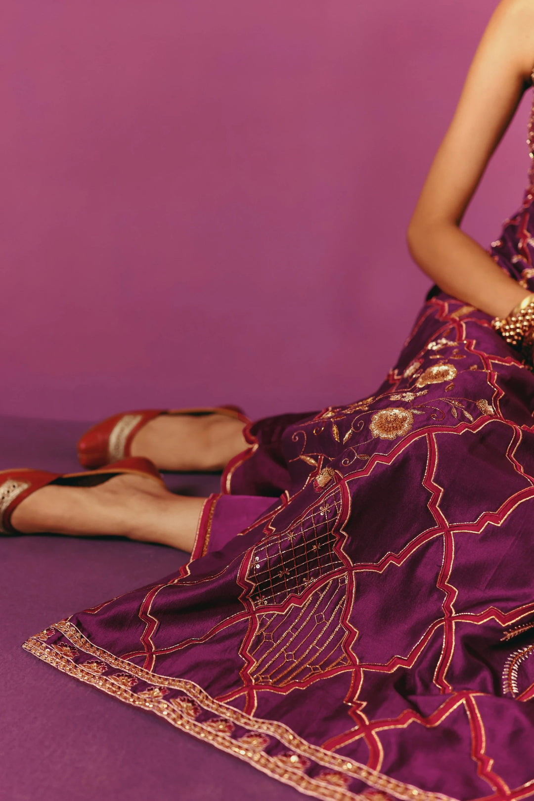 Close-up of the hem of Warra's wine-purple kurta showing gold quatrefoil trellis embroidery, worn with red block-heel juttis