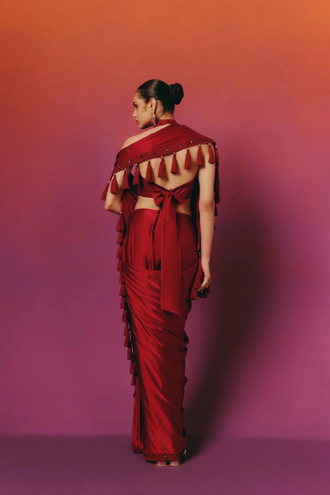 Back view of a model in Warra's crimson saree showing the beaded tassel trim over one shoulder, a bow-tied open-back blouse and gold jewellery
