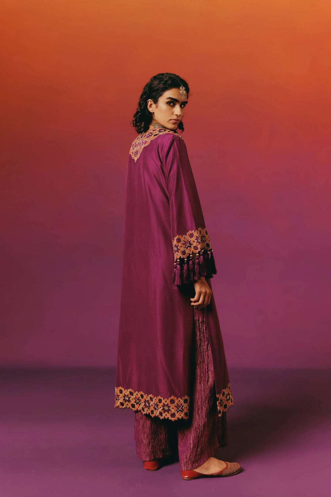 Back view of a model in Warra's magenta silk kaftan showing gold-and-orange star-lattice embroidery on the yoke and tasselled sleeve cuffs, worn with matching shimmer palazzo pants