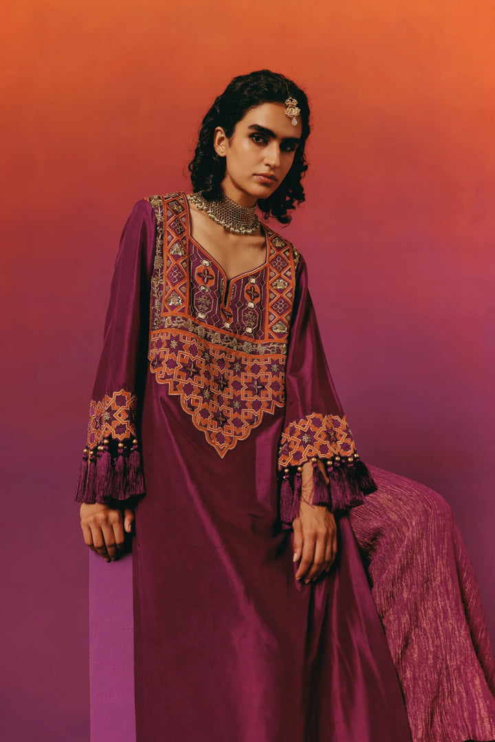 Full-length seated three-quarter view of a model in Warra's magenta silk kaftan showing the tasselled, star-embroidered sleeve draped over the shimmer palazzo pants