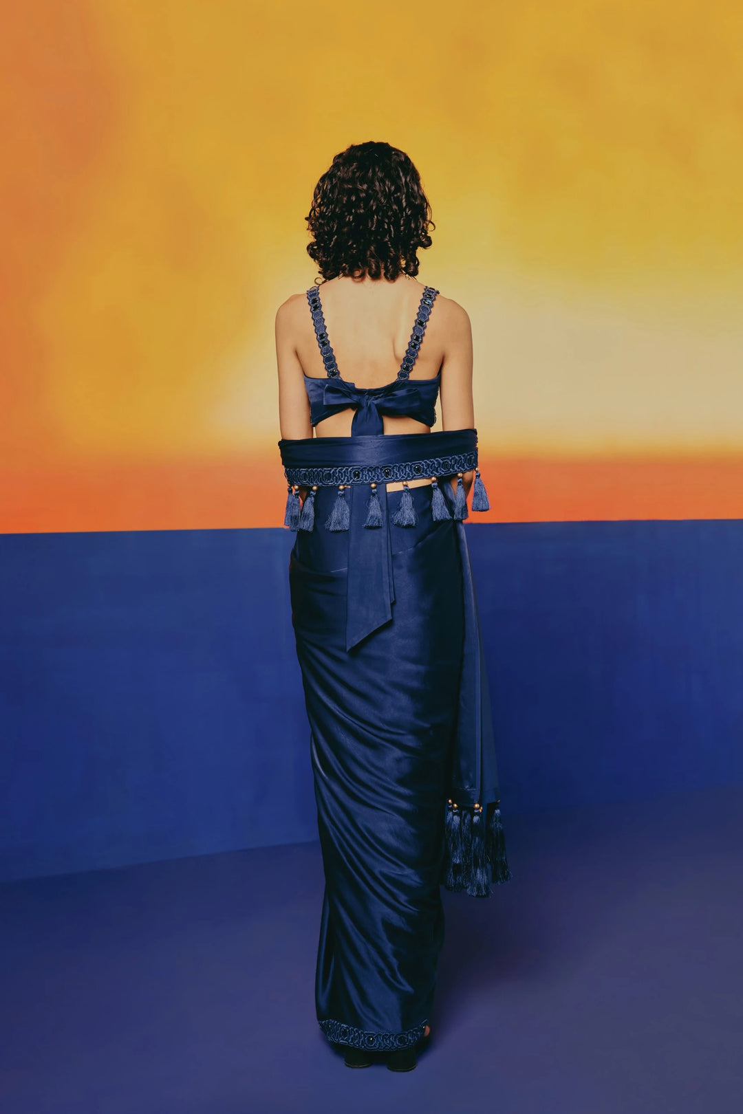 Back view of a model in Warra's Crystal Blue Tassel Saree showing the bow-tied blouse back and tasseled waist sash over a sheath skirt