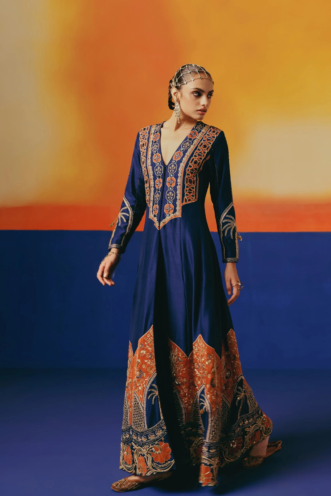 Full-length front view of a model walking in Warra's navy silk anarkali with an orange-and-gold jaal embroidered yoke and a palm tree and floral hem border, paired with a gold mathapatti