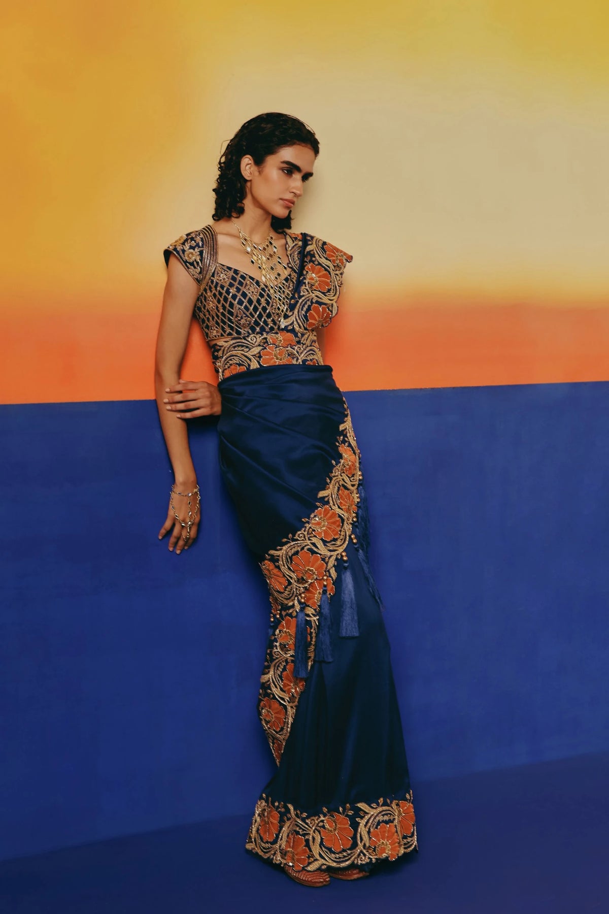 Three-quarter front view of a model leaning against a wall in Warra's navy silk saree with orange floral embroidered pallu