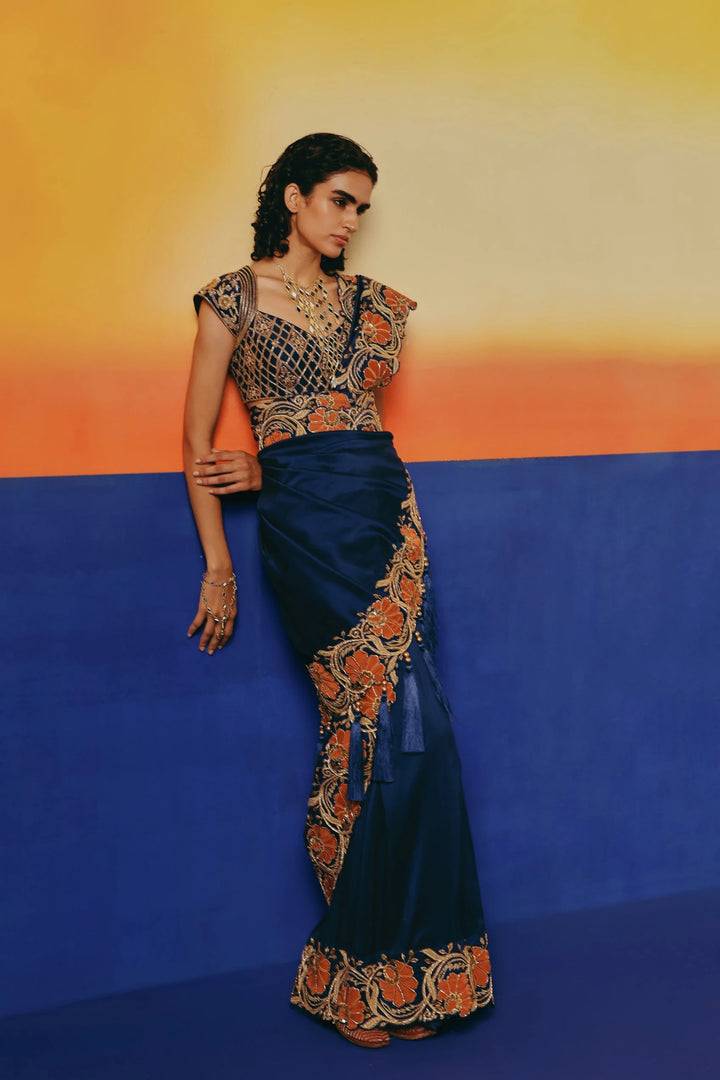 Three-quarter front view of a model leaning against a wall in Warra's navy silk saree with orange floral embroidered pallu
