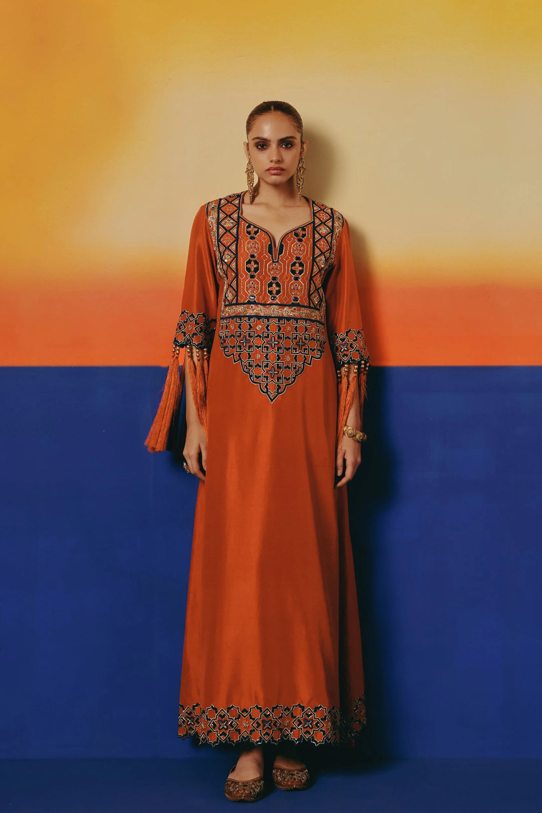 Full-length front view of a model in Warra's rust-orange silk kaftan with tasselled sleeve cuffs and an embroidered hem border