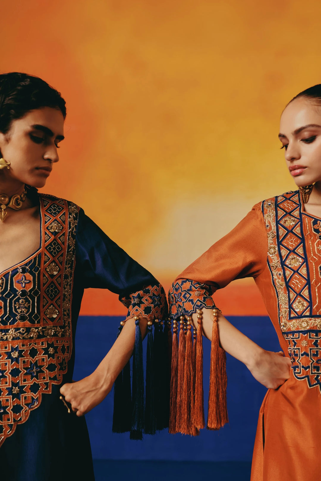 Two models  in Warra's rust-orange silk kaftan with gold lattice embroidery and tasselled sleeve fringe