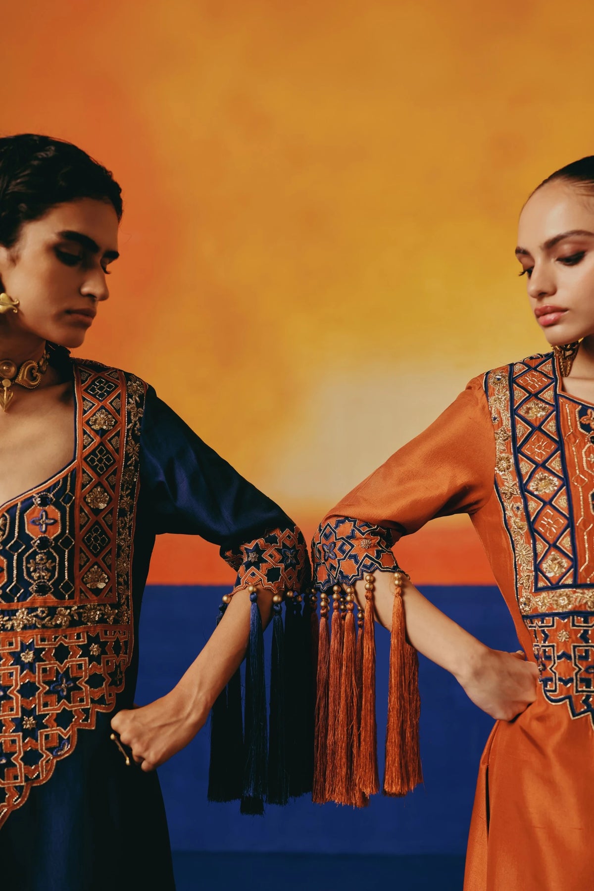 Two models  in Warra's rust-orange silk kaftan with gold lattice embroidery and tasselled sleeve fringe