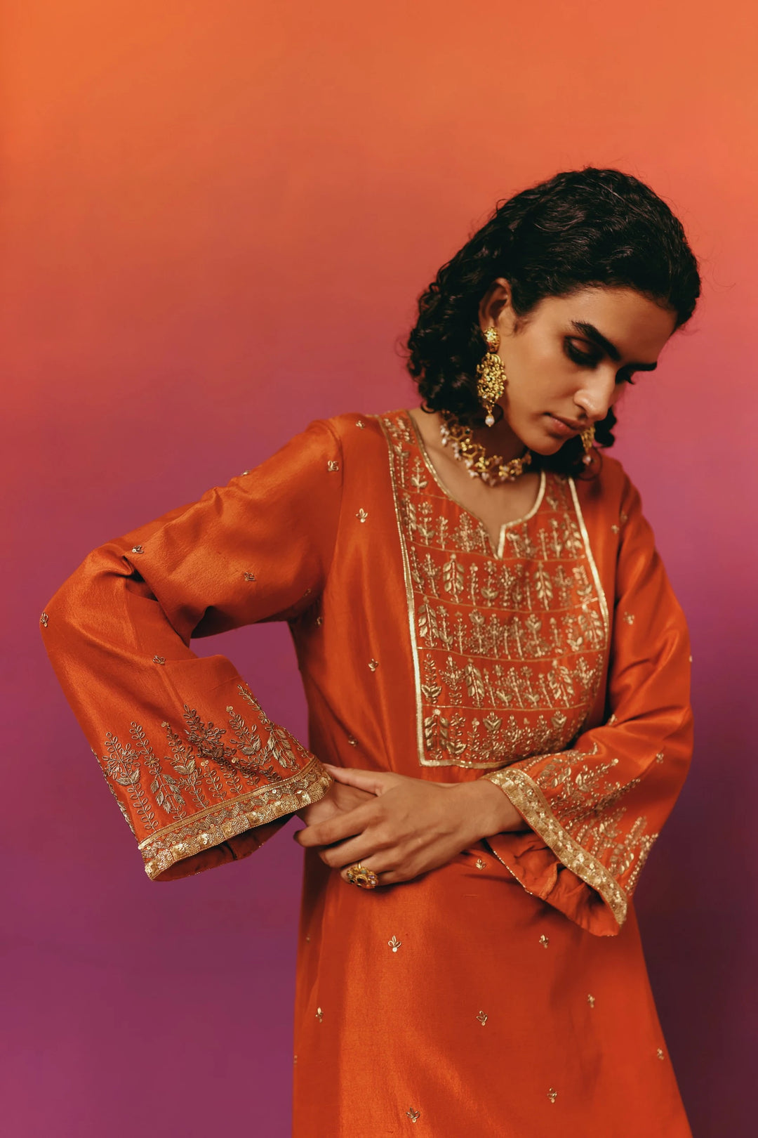 Close-up of a model in Warra's orange silk kurta showing a gold embroidered floral yoke panel in horizontal rows, gold jhumka earrings and a layered necklace, hand on hip