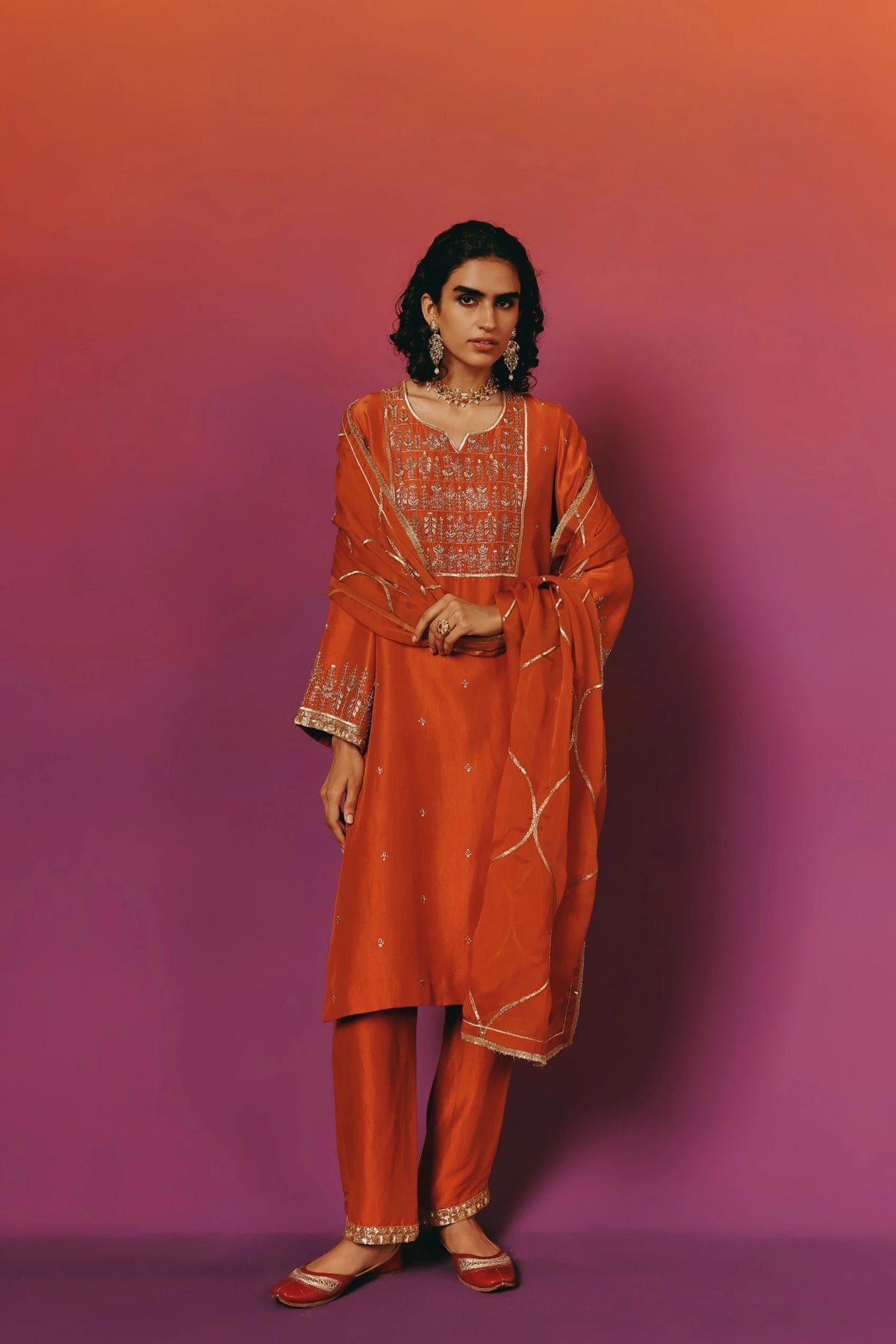 Full-length front view of a model in Warra's orange silk kurta with a gold embroidered floral yoke, gold-trimmed dupatta with wavy vine embroidery draped over one arm, worn with matching pants