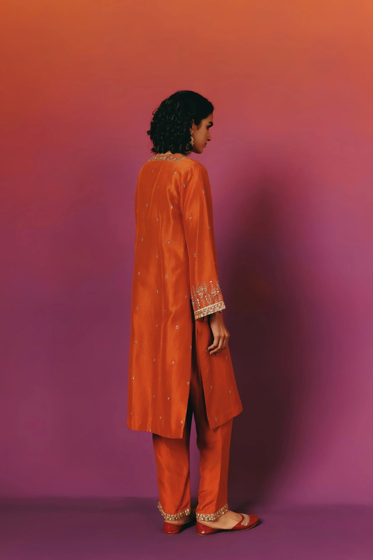 Full-length back/side view of a model in Warra's orange silk kurta with gold embroidered floral cuffs and scattered gold floret dots, worn with matching wide-leg pants and red juttis
