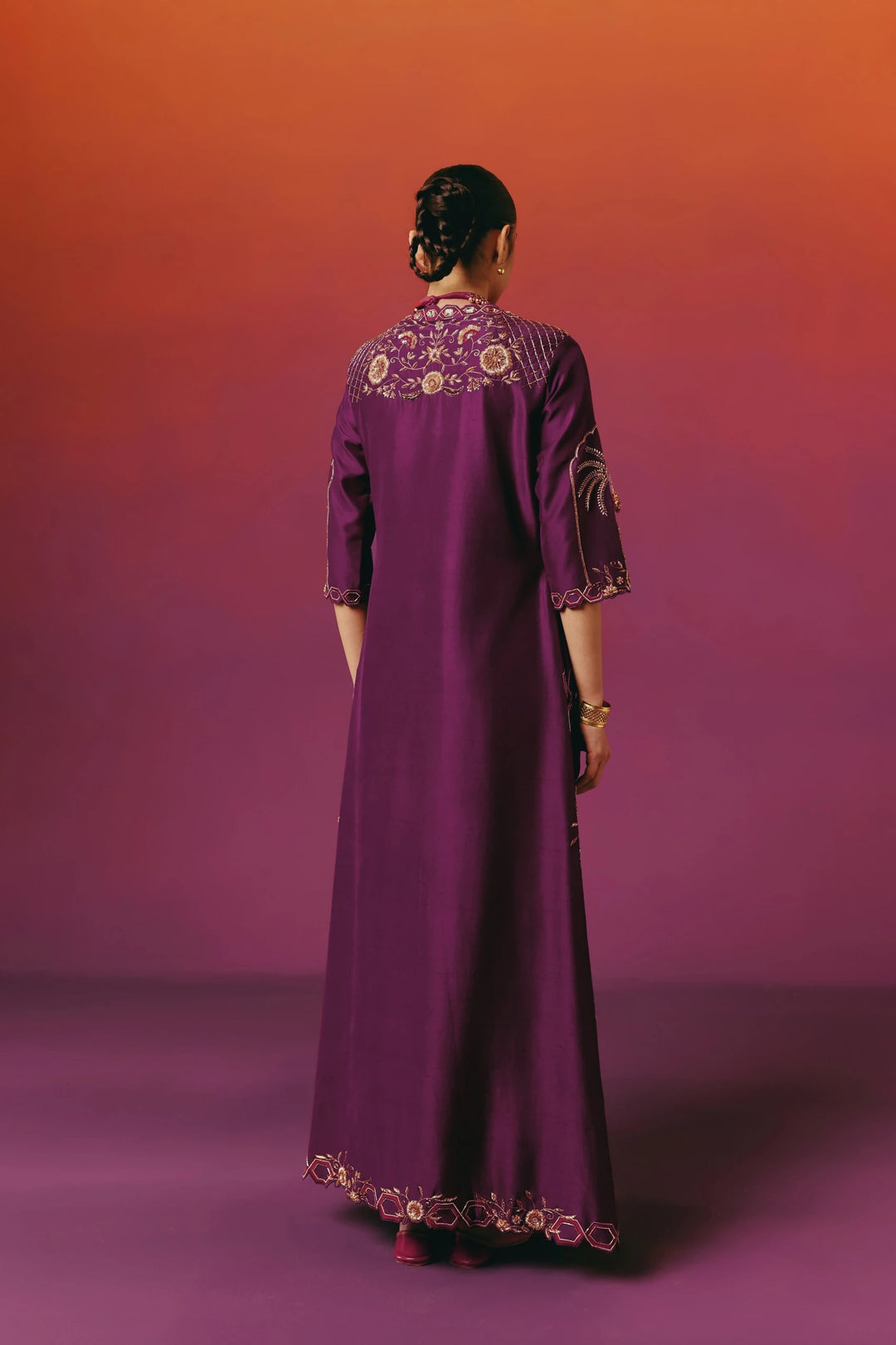 Back view of a model in Warra's deep purple silk kaftan, full length, with gold floral embroidered yoke on the back panel, matching embroidered sleeve cuffs and a hexagon-bordered floral hem