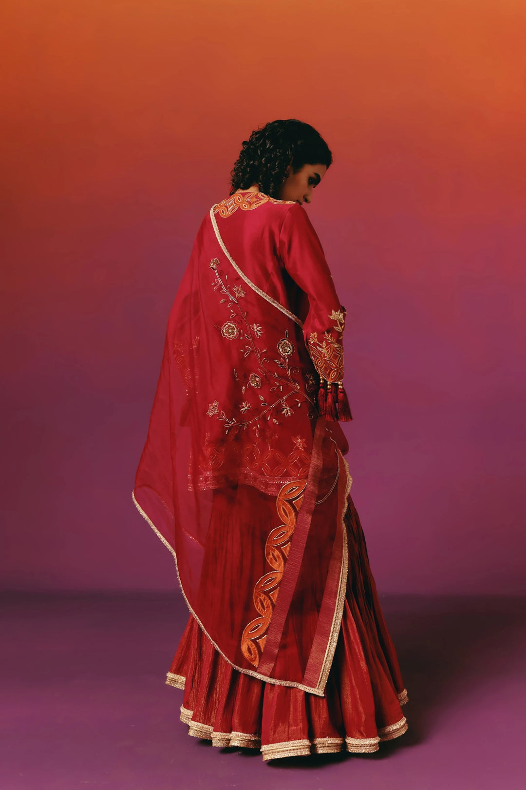 Two models seated together, one in Warra's red-and-orange embroidered sharara set and one in a purple saree, showing matching gold jewellery and tasselled sleeve details