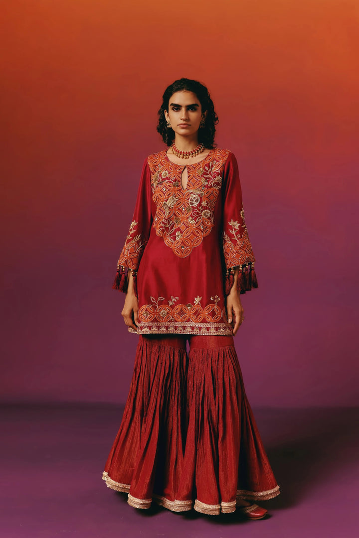 Full-length front view of a model in Warra's deep red silk kurta with an orange embroidered yoke in a circular medallion pattern, paired with flared sharara pants and a gold choker