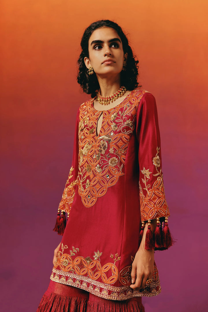 Close-up front view of a model in Warra's red silk kurta showing the orange embroidered circular medallion yoke and tasselled sleeve cuffs, worn over sharara pants