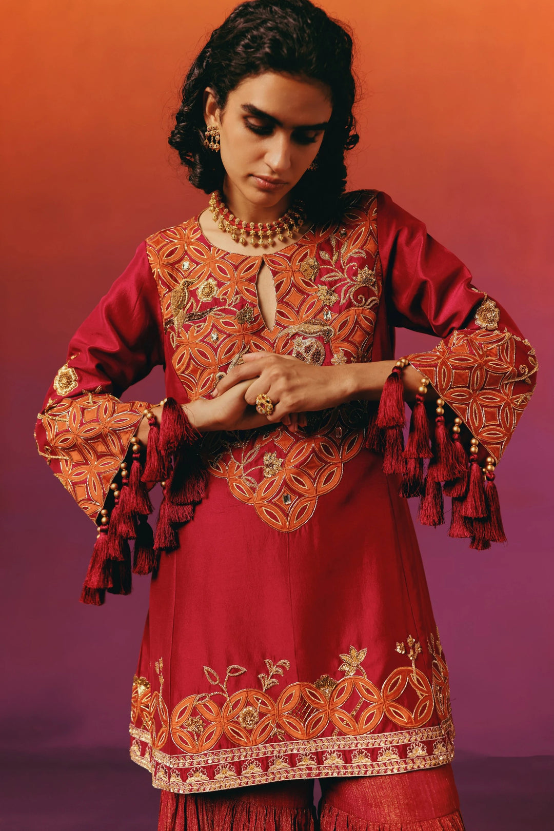 Close-up of Warra's red silk kurta with hands clasped at the waist, showing the tasselled sleeve cuffs and orange-embroidered medallion yoke in motion