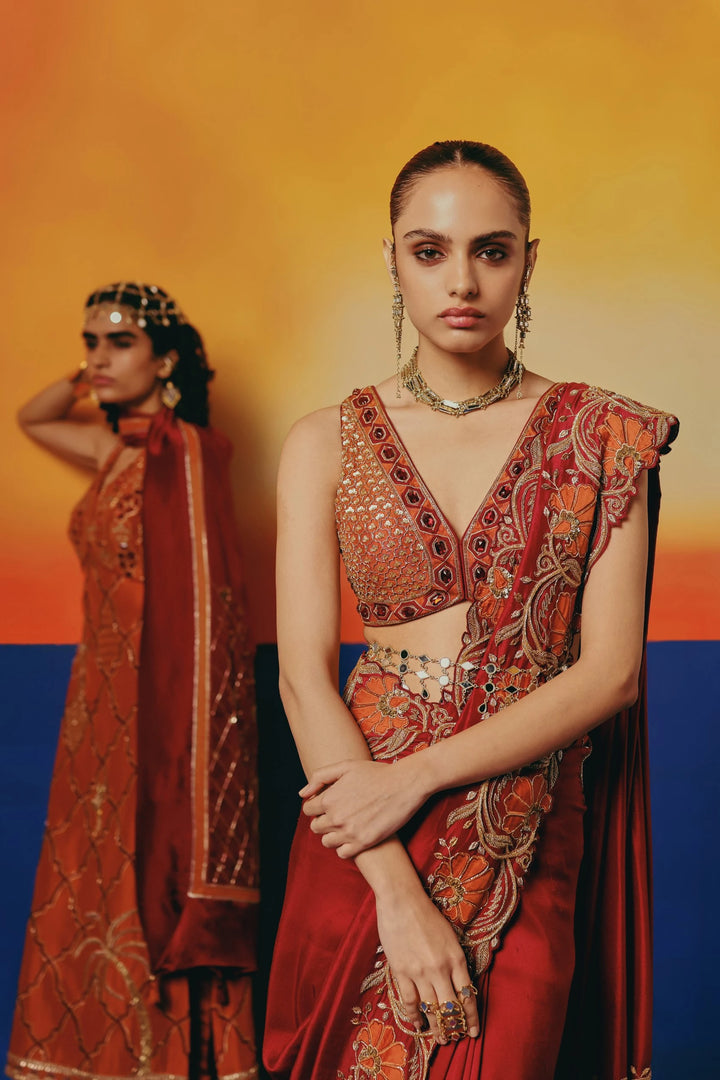 Close-up portrait of a model in Warra's red silk saree with orange floral embroidery and layered gold jewellery, orange kurta set blurred behind