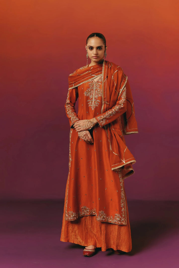 Full-length front view of a model in Warra's rust-orange silk kurta with gold floral embroidery on the yoke and sleeves, paired with a matching embroidered dupatta, sharara, and pearl hath-phool hand jewelry